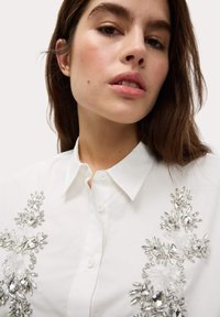 Camicia bianca con decorazioni in cristallo disposte in motivi floreali sul davanti. Colletto classico e chiusura a bottoni visibili.