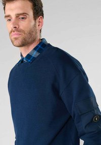 Maglione blu navy con texture a costine e colletto rotondo, indossato sopra una camicia a quadri blu con colletto. Presenta una tasca a patta sulla manica.