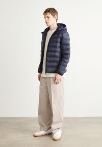 Giubbotto trapuntato blu navy con cappuccio, chiusura a zip e design trapuntato. Indossato sopra un top beige chiaro e abbinato a pantaloni beige larghi e sneakers bianche.