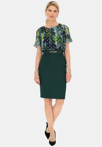 Bluzka w kwiaty w kolorze granatowym, zielonym i niebieskim; w komplecie z ciemnozieloną spódnicą o kroju ołówkowym z szerokim paskiem w talii, co zapewnia dopasowany krój.