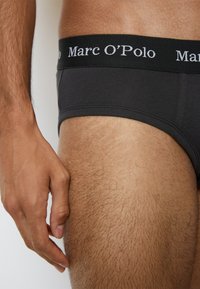 Svarta bomullsunderkläder med en mjuk midja med "Marc O'Polo"-logotyp. Närbild av hand som vilar på benet, vilket visar hårtexturen.
