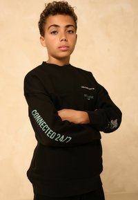 Schwarzer Sweatshirt mit langen Ärmeln, ausgestattet mit grünem Text "CONNECTED 24/7" auf einem Arm und verschiedenen Druckdetails auf der Vorderseite und dem Ärmel.
