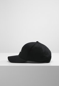 Svart bomullsbasketcaps med buet visir, med en hvit brodert logo på framsiden og seks ventilasjonsøyler.