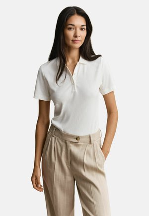 Femme aux longs cheveux foncés portant un polo côtelé blanc à manches courtes, rentré dans un pantalon plissé beige taille haute avec poches.