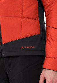 Veste isolée rouge et noire avec texture matelassée, présentant une fermeture éclair et un logo Vaude bien visible en bas à gauche.