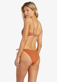 Žena s blond vlasmi, oblečená v bikiny v hnedej farbe, stojí s chrbtom a hlavou otočenou k fotoaparátu na bielom pozadí.