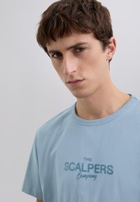 Joven con el cabello corto y marrón que lleva una camiseta de color azul claro con el logo de "THE SCALPERS Company", mirando seriamente a la cámara frente a un fondo sencillo.