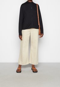Sort langærmet skjorte med knapper, lys beige brede bukser og brune sandaler med flad sål. En brun crossbody taske fuldender looket.
