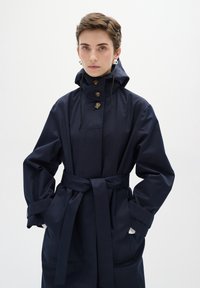 Jūras zila ūdensizturīga antitrešs ar augstu apkakli, slēdzamiem pogām, regulējamu auklu un sasietu jostas jostu. Redzamas garas piedurknes un sānu kabatas.