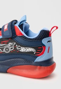 Sinine ja tumesinine tennisetoss, millel on punased aktsendid, võrgumuster ja Hot Wheels graafika. Velcro rihm ja tõmbeava mugavaks selga panemiseks.
