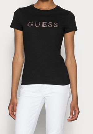 Persoon in een zwart T-shirt met korte mouwen met glinsterende "GUESS" tekst en witte broek, staand tegen een effen lichte achtergrond.