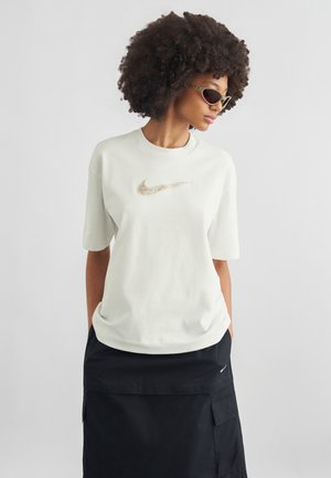 Γυναίκα με πλούσια σγουρά μαλλιά που φοράει γυαλιά ηλίου με σχήμα γάτας, λευκό t-shirt Nike και μαύρη φούστα με τσέπες, στέκεται μπροστά σε ένα απλό φόντο.