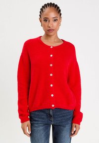 Ung kvinde med fletninger iført en lys rød knaptrøje og blå jeans, stående mod en ensfarvet lys baggrund.