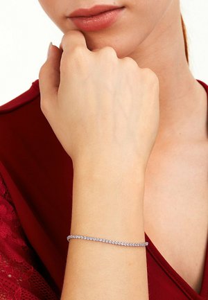 Nahaufnahme eines Handgelenks mit einem dünnen Silberarmband aus kleinen verbundenen Steinen, getragen von einer Person in einem roten Top mit Spitzenärmeln.