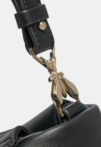 Tracolla di una borsa in pelle nera con un luccicante gancio in metallo dorato che presenta un design di figura astratta, fissata da un anello in pelle con un bottoncino.