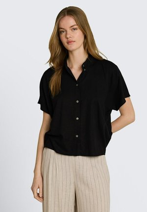 Młoda kobieta z długimi włosami, ubrana w luźną czarną koszulę z krótkim rękawem na guziki i beżowe spodnie w cienkie paski, stoi z jedną ręką za plecami.