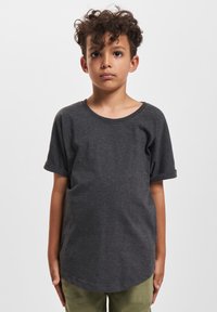 Dreng med krøllet hår står med ansigtet fremad iført en mørkegrå T-shirt med korte ærmer og olivengrønne bukser foran en ensfarvet hvid baggrund.