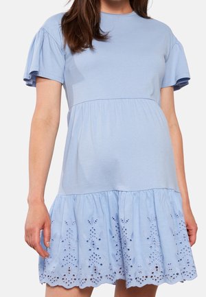 Mujer con un vestido azul claro de manga corta, con borde fruncido y detalles de bordado calado en la parte inferior.