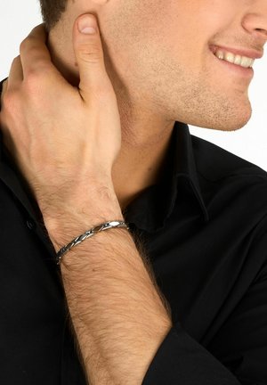 Uomo sorridente che indossa una camicia nera e un braccialetto a catena d'argento, con la mano che tocca il lato del collo.