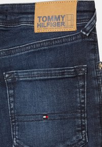 Jeans de mezclilla azul oscuro con un acabado texturizado, que presentan una etiqueta de cuero marrón y un pequeño logotipo tricolor en el bolsillo trasero.