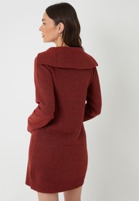 Vrouw met lang bruin haar, gekleed in een roestkleurige gebreide jurk met een brede kraag, poseert met het gezicht naar links tegen een witte achtergrond.