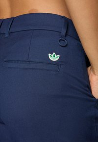 Dettaglio ravvicinato di pantaloni blu navy con una tasca posteriore, un piccolo logo a foglia verde e bianca e un anello metallico sul cinturino.