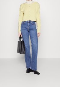 Cardigan giallo lavorato a maglia con motivo a rombi, con bottoni frontali, abbinato a jeans blu a vita alta e borsa in pelle nera. Scarpe nere a punta.