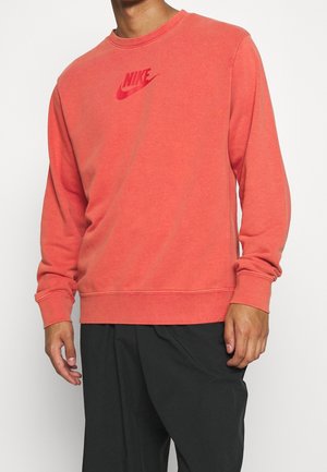 Man draagt een koraalrode Nike-trui met lange mouwen en zwarte broek tegen een witte achtergrond.