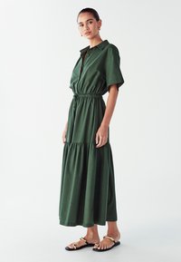 Abito verde scuro a maniche corte con chiusura a bottoni e vita elastica. Gonna a strati fluida, realizzata in tessuto morbido. Indossato con sandali neri.