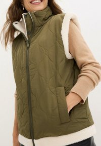 Gilet trapuntato verde oliva con colletto alto e rifiniture in pile crema, dotato di una zip frontale e tasche laterali. Texture morbida, design senza maniche.