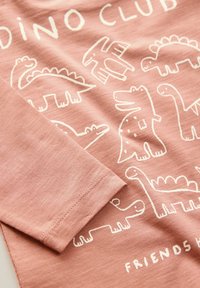 Světle růžové tričko s bílými ilustracemi dinosaurů a textem "DINO CLUB". Krátký rukáv, texturovaná tkanina, volný střih, neformální design.