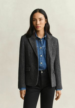 Vrouw met lang donker haar die een grijze visgraatblazer draagt over een blauw denim overhemd en zwarte broek, staand tegen een effen lichte achtergrond.