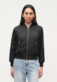 Μαύρο μπουφάν bomber με χρυσές λεπτομέρειες φερμουάρ, ριμπ μανσέτες και τελείωμα. Διαθέτει κεντημένο λογότυπο στο αριστερό στήθος. Ομαλή υφή υφάσματος.