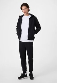 Svart zip-up hoodie, vit T-shirt, svarta mjukisbyxor och svarta sneakers. Tyget är mjukt och har minimal märkning på bröst och ben.