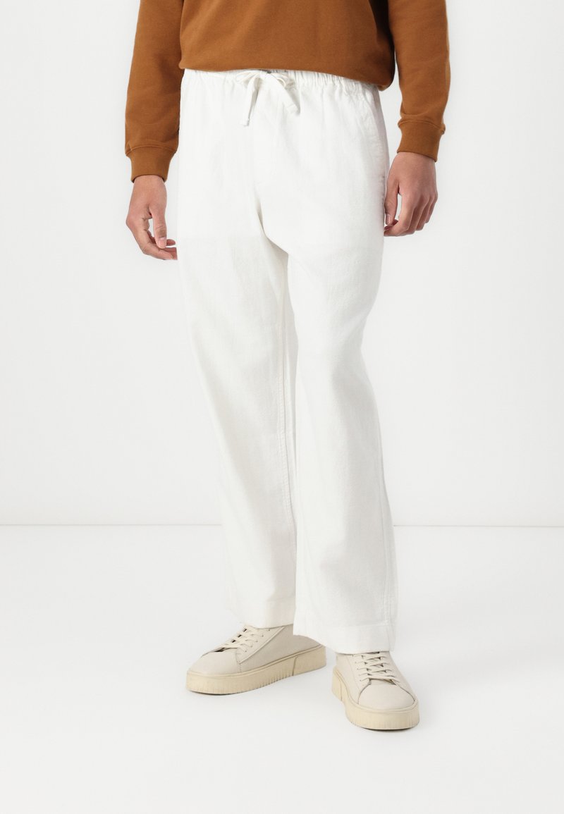 Pantalones holgados blancos con cintura elástica y cordón, hechos de tejido texturizado, combinados con zapatillas de deporte color blanco roto.