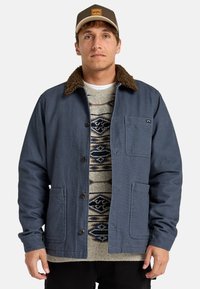 Navy blue jacket with a brown fleece collar, button closure, and front pockets, layered over a patterned sweater and paired with dark pants.