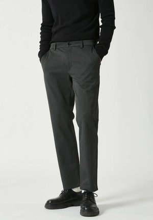 Mann trägt dunkelgraue maßgeschneiderte Hose, schwarzen Pullover, weiße Socken und schwarze Schnürschuhe, steht mit den Händen in den Taschen.