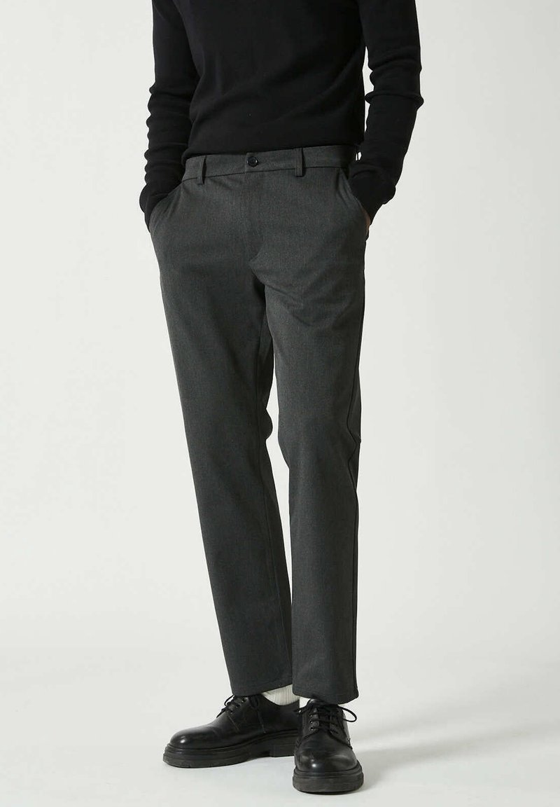 Homme portant un pantalon gris foncé sur mesure, un pull noir, des chaussettes blanches et des chaussures à lacets noires, debout les mains dans les poches.