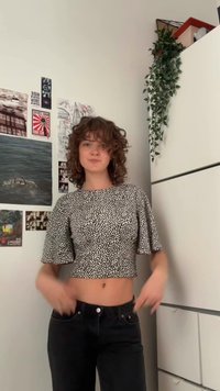 Melnā un baltā kratītā topa daļa ar punktiņainu rakstu, īsām izplestām piedurknēm, kombinēta ar augsto vidukli melnām biksēm, fons ir neitrāla siena.