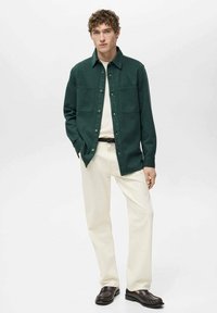 Giacca camicia verde scuro con due tasche sul petto, chiusura a bottone, indossata su un maglione crema e pantaloni bianchi, con scarpe slip-on marrone scuro.