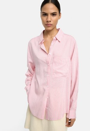 Kobieta ubrana w luźną różowo-białą koszulę w paski zapinaną na guziki z przednią kieszenią, w połączeniu z jasną, prążkowaną spódnicą.