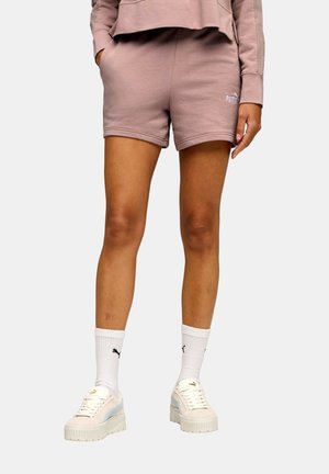 Osoba oblečená v purpurových šortkách Puma a zodpovedajúcej mikine, biele ponožky Puma a krémové tenisky na platforme, stojaca s rukami v náprsných vreckách.