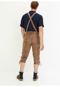 Pettorina in suede marrone con bretelle regolabili, di lunghezza cropped e lacci laterali. Indossata sopra una maglietta scura e abbinata a stivali neri.