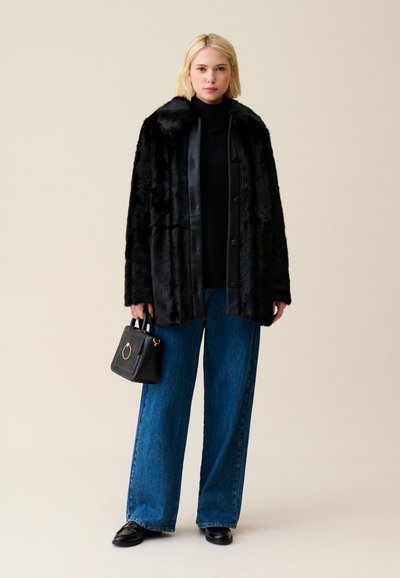 Cappotto in pelliccia sintetica nera con colletto in raso, bottoni frontali e jeans larghi in denim blu. Il modello porta una piccola borsa nera con un dettaglio dorato.