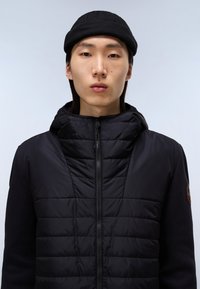 Černá bunda s prošívaným předním dílem, kapucí a kontrastními pletenými rukávy. Zapínání na zip a logo nášivka na ruce. Hladký materiál a přiléhavý střih.