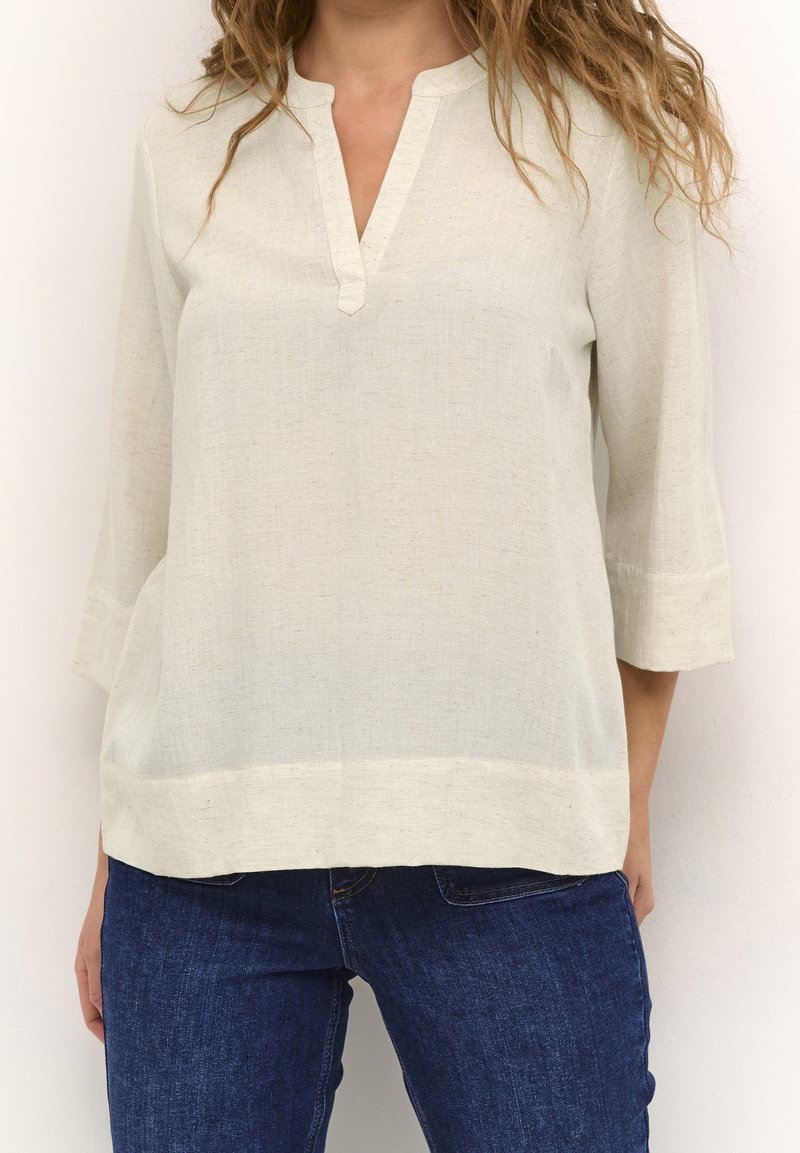 Femme portant un chemisier ample à manches longues beige clair avec une encolure fendue et un jean bleu foncé, debout devant un fond uni.