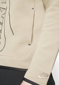Beige Sweatshirt mit schwarzen Akzenten und gesticktem Nike-Logo am Bündchen. Verfügt über eine glatte Textur und ein schlankes Design mit versteckten Taschen.