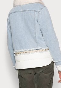 Vaaleansininen farkkutakki, jossa on valkoinen toppaushelma ja kukkakoristeet. Siinä on repsottavat reunat ja keinoturkiskaulus.