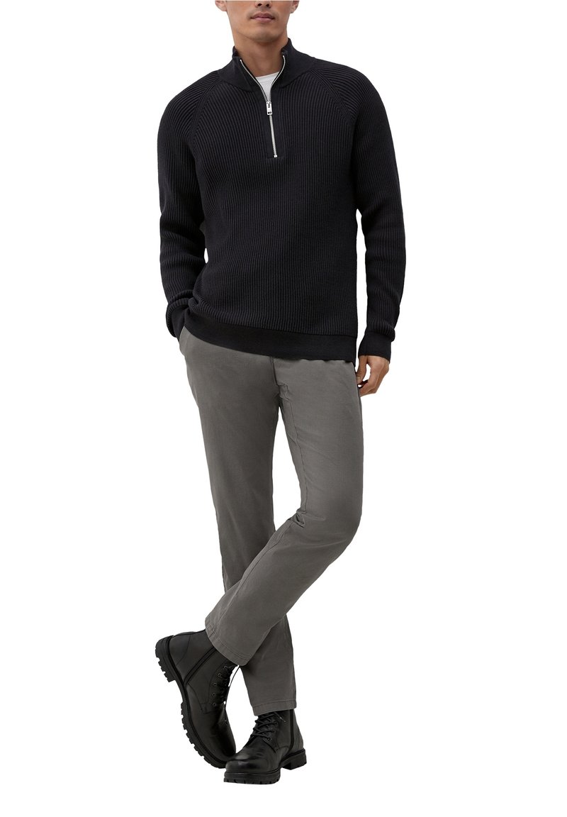 Mies, jolla on musta ribbineulottu puolivetoketjullinen neulepaita, harmaat housut ja mustat nyörisaappaat, seisoo ristissä jalat ja kädet taskuissa.