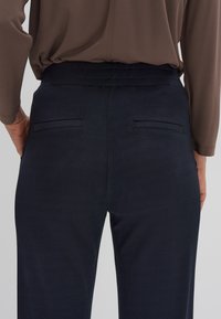 Marineblauwe broek van een gestructureerde stof, met twee achterzakken en een gladde tailleband, gezien van de achterkant.